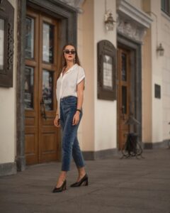 Fashionable woman in sunglasses and jeans poses by ornate wooden doors.