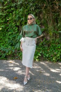 Fashionable woman wearing striped top and skirt poses outdoors with lush greenery backdrop.