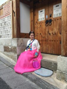 East Asian woman in vibrant hanbok, sitting by a traditional doorway in Seoul's Gahoe-dong.