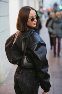 Trendy woman in black leather jacket poses confidently on an urban street with blurred pedestrians.
