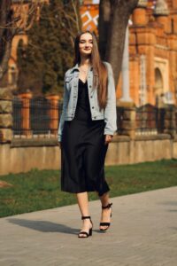 Fashionable woman in denim jacket and black dress walking along a sunny sidewalk.