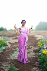 A joyful woman wearing a lavender saree stands outdoors, flashing peace signs amid nature's beauty.