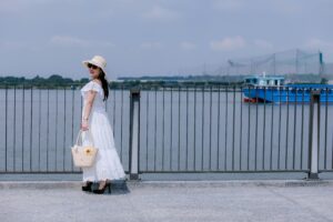 Fashionable woman in white dress by a waterfront with a blue boat passing by.