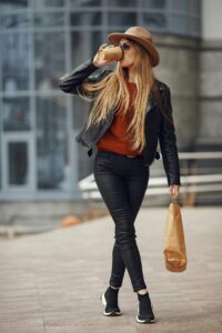 Stylish woman drinking coffee while walking on the street, wearing leather jacket and sunglasses.