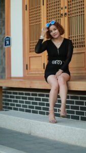 Fashionable woman sitting on steps wearing a black dress, accessorized with jewelry, posing stylishly outdoors.